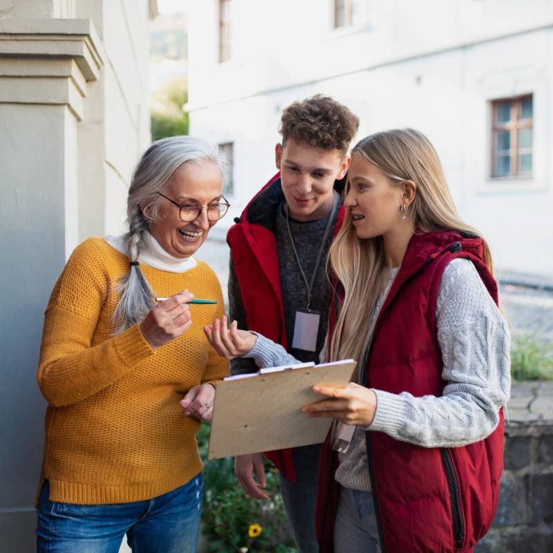 Young door to door volunteers talking to senior woman and taking a survey at her front door.